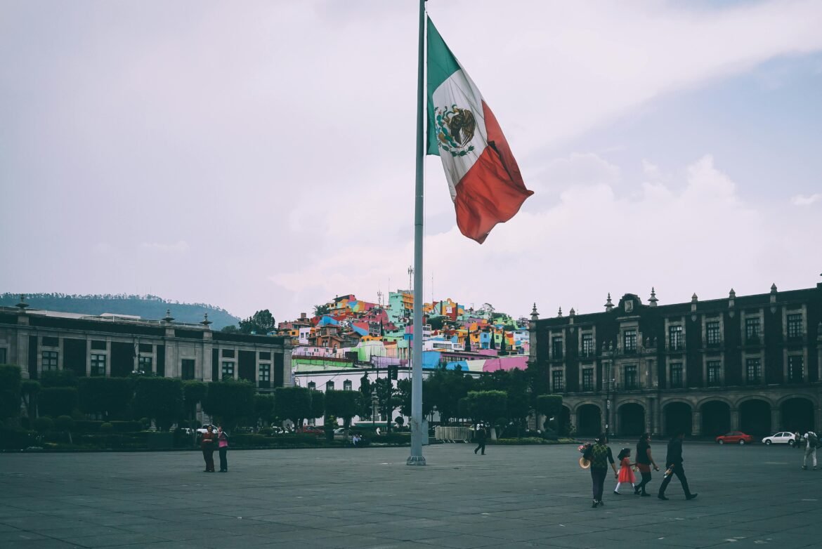Large Mexican flag waving in a city plaza with colorful hillside houses in the background and people walking in the foreground.