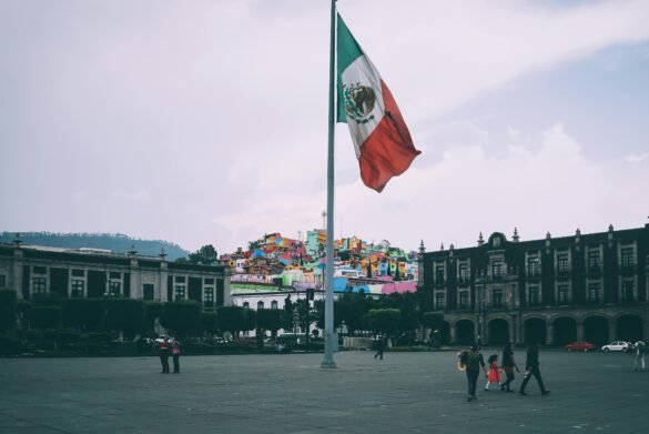 Large Mexican flag waving in a city plaza with colorful hillside houses in the background and people walking in the foreground.